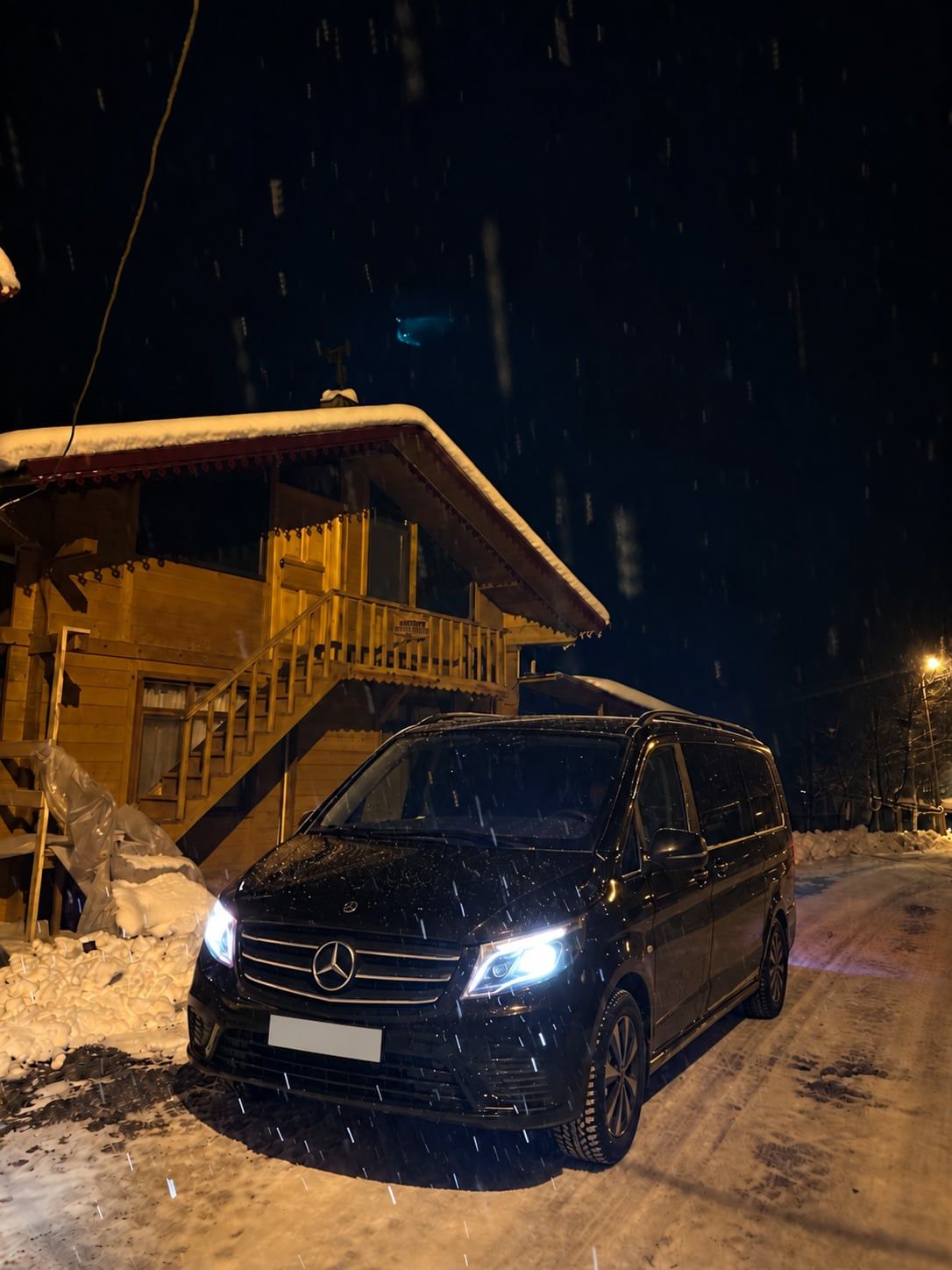 Kar yağışı altında ahşap yayla evinin önünde farları açık siyah Mercedes Vito